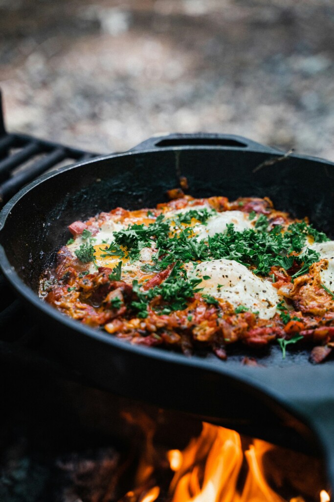 A delicious campfire-cooked shakshuka-style meal in a cast iron skillet, topped with eggs and fresh herbs—perfect for those looking for easy camping meals and the best camping meals ideas.