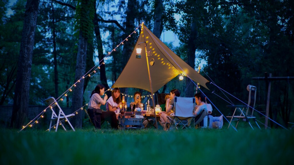 A group of campers enjoying an evening under a beautifully lit canopy in a forest, surrounded by fairy lights and camping gear—capturing the perfect vibe for an Electric Forest Camping Guide.