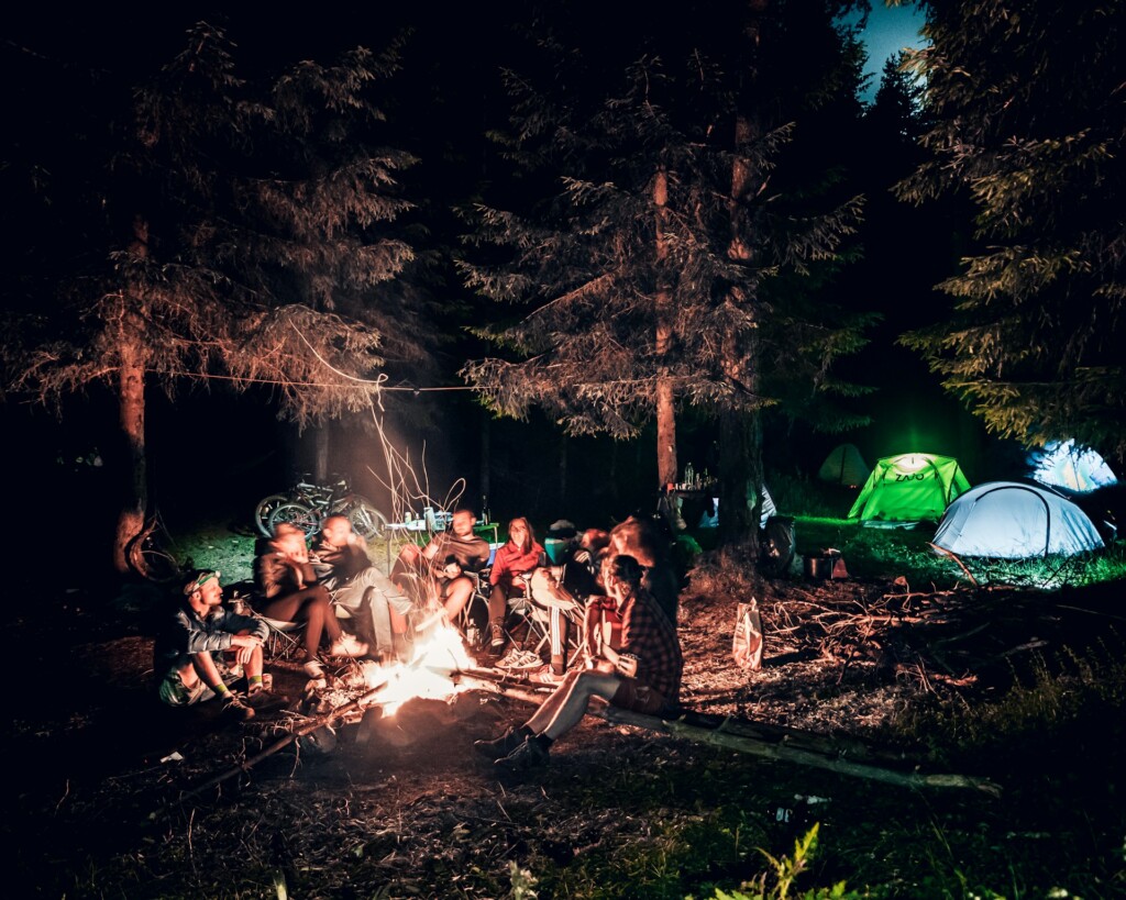A group of friends gathered around a campfire at night, sharing stories, laughing, and enjoying their camping experience in the woods.
